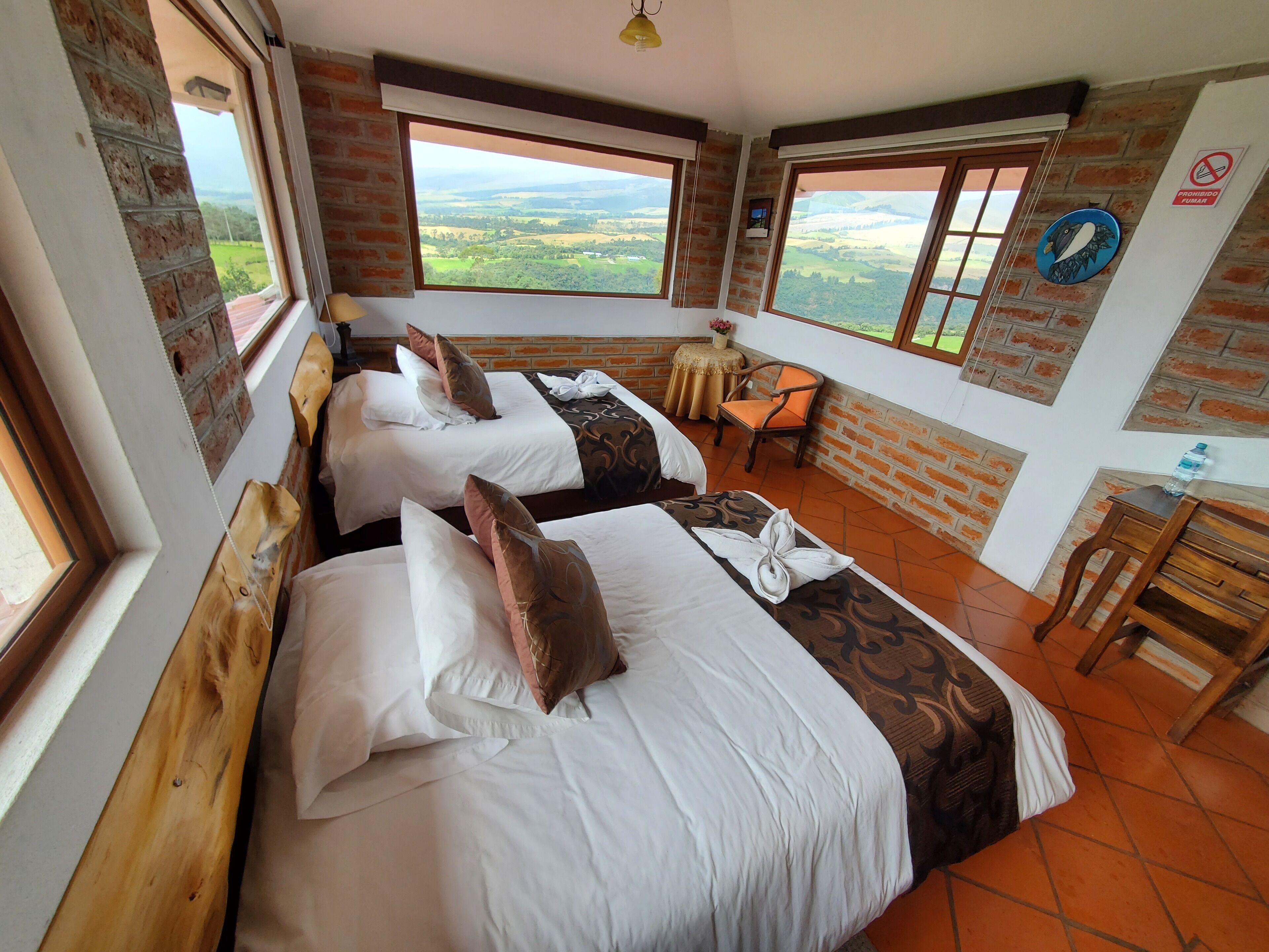 Habitación de hotel con dos camas, una mesita y una silla. Amplios ventanales con vistas al campo.