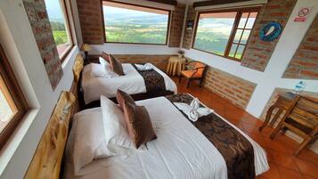 Habitación de hotel con dos camas, una mesita y una silla. Amplios ventanales con vistas al campo.