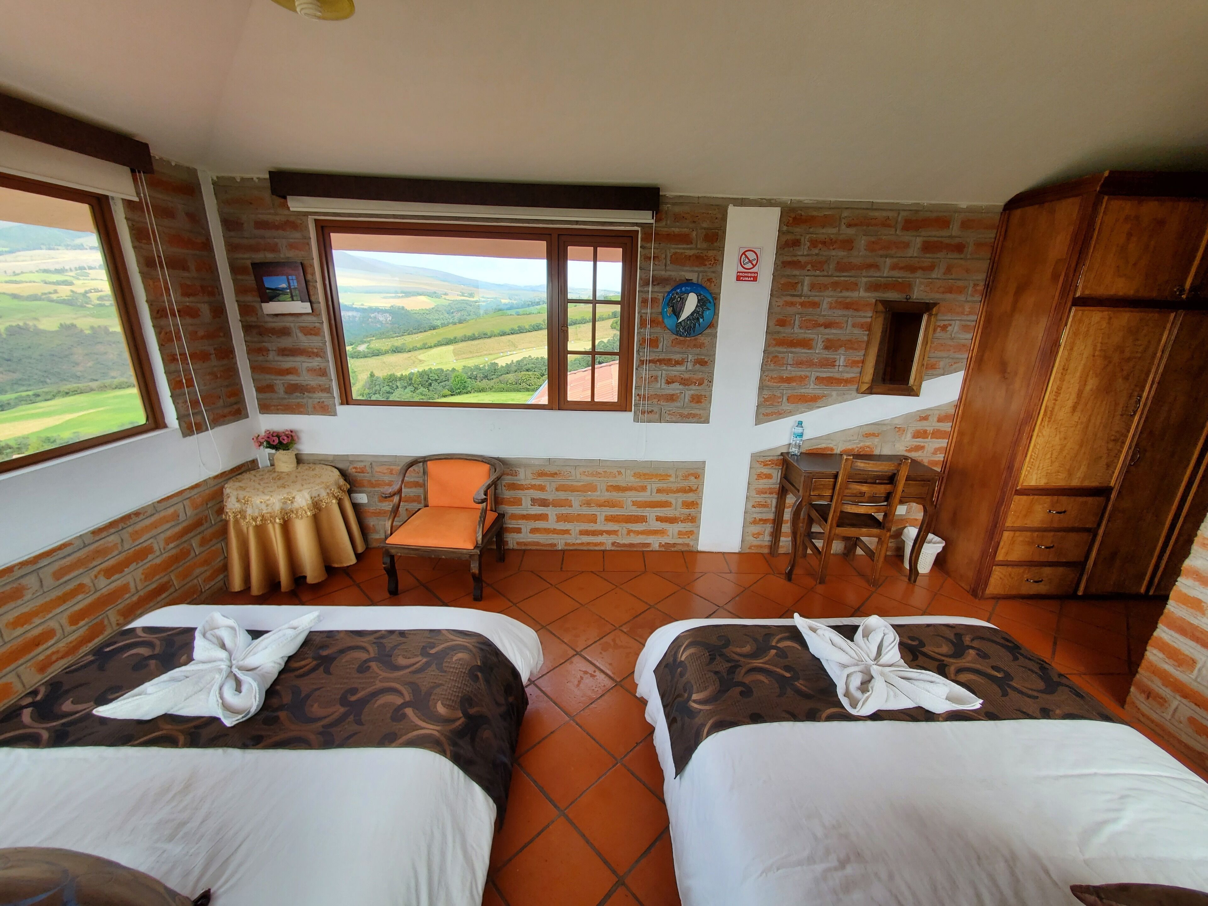 A room with two beds, a small table with a chair, a wooden cabinet, and a window with a view of the countryside.
