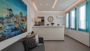 A hotel lobby with a reception desk, a large painting of a coastal town, a clock, and a sofa with decorative pillows. - Nikolas Hotel (Santorini)