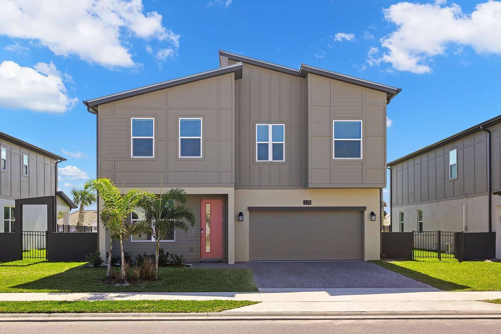 A two-story residential building with a garage, a red door, and a small front yard with a palm tree.