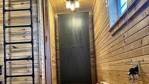 A narrow hallway with wooden walls and a door at the end. A ladder is leaning against the wall on the left, and a heater is mounted on the wall to the right.