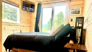 A wooden cabin bedroom with a bed, a nightstand, a TV mounted on the wall, and a window with curtains.