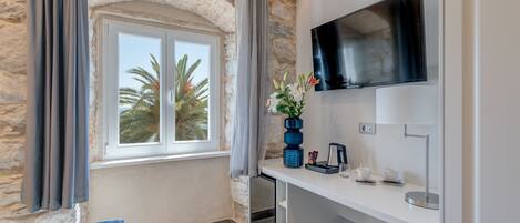 A room with a stone wall, a window with a view of palm trees, a flat-screen TV mounted on the wall, a white desk with a vase of flowers, and a bed with blue bedding.
