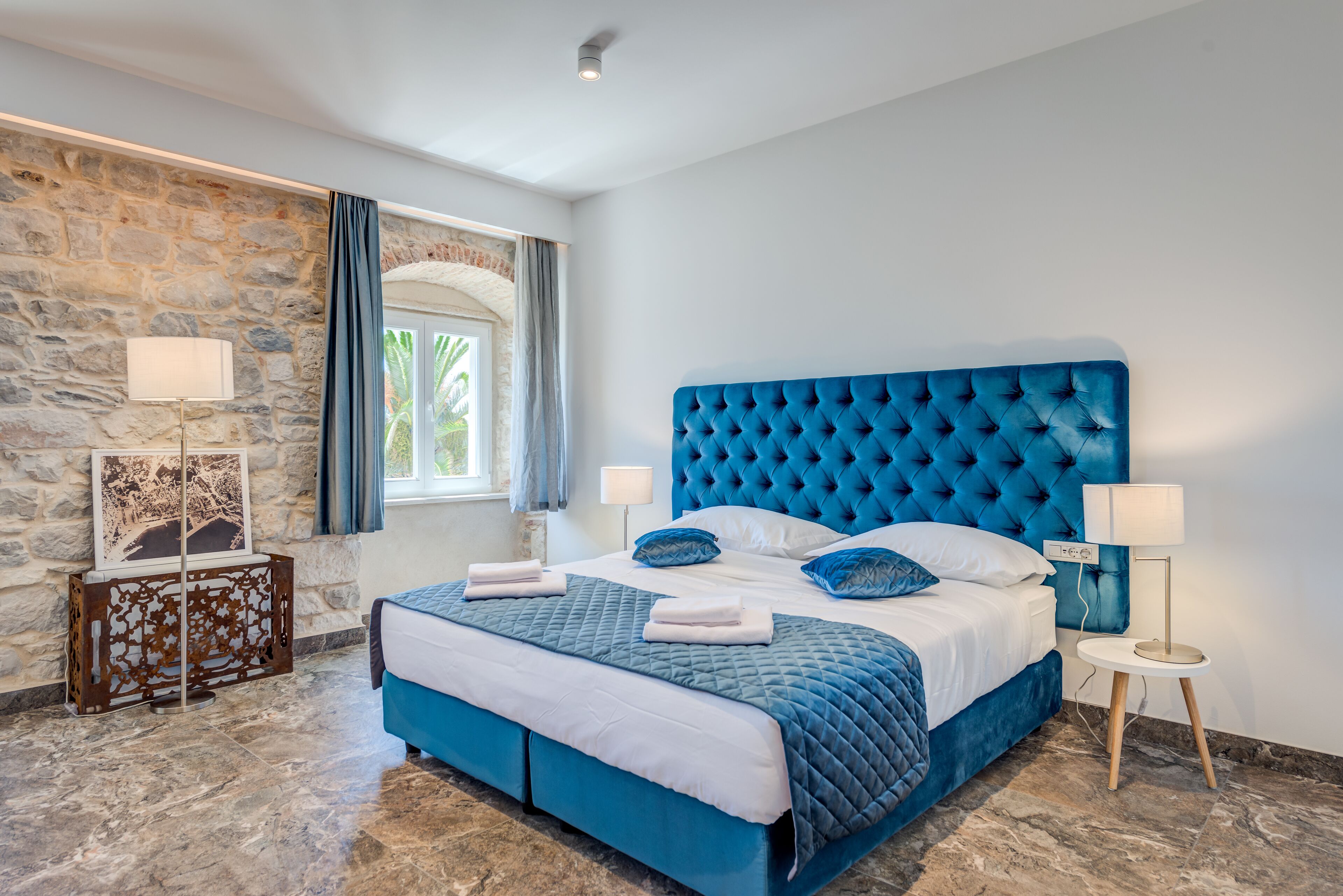 A bedroom with a stone wall, a large bed with blue bedding, and two bedside lamps.