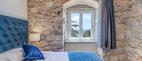 A bedroom with a stone wall, a window with curtains, a bed with blue bedding, and a white headboard.