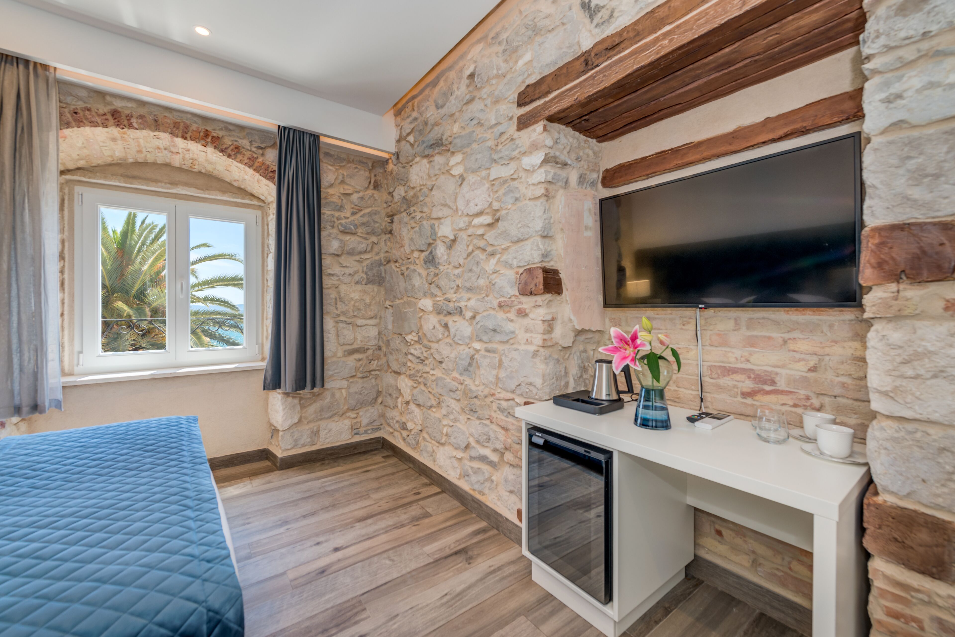 Una stanza con parete in pietra, una TV a schermo piatto montata, soffitto in legno, un letto con copriletto blu, un tavolino con una macchina per caffè e una finestra vista palme.