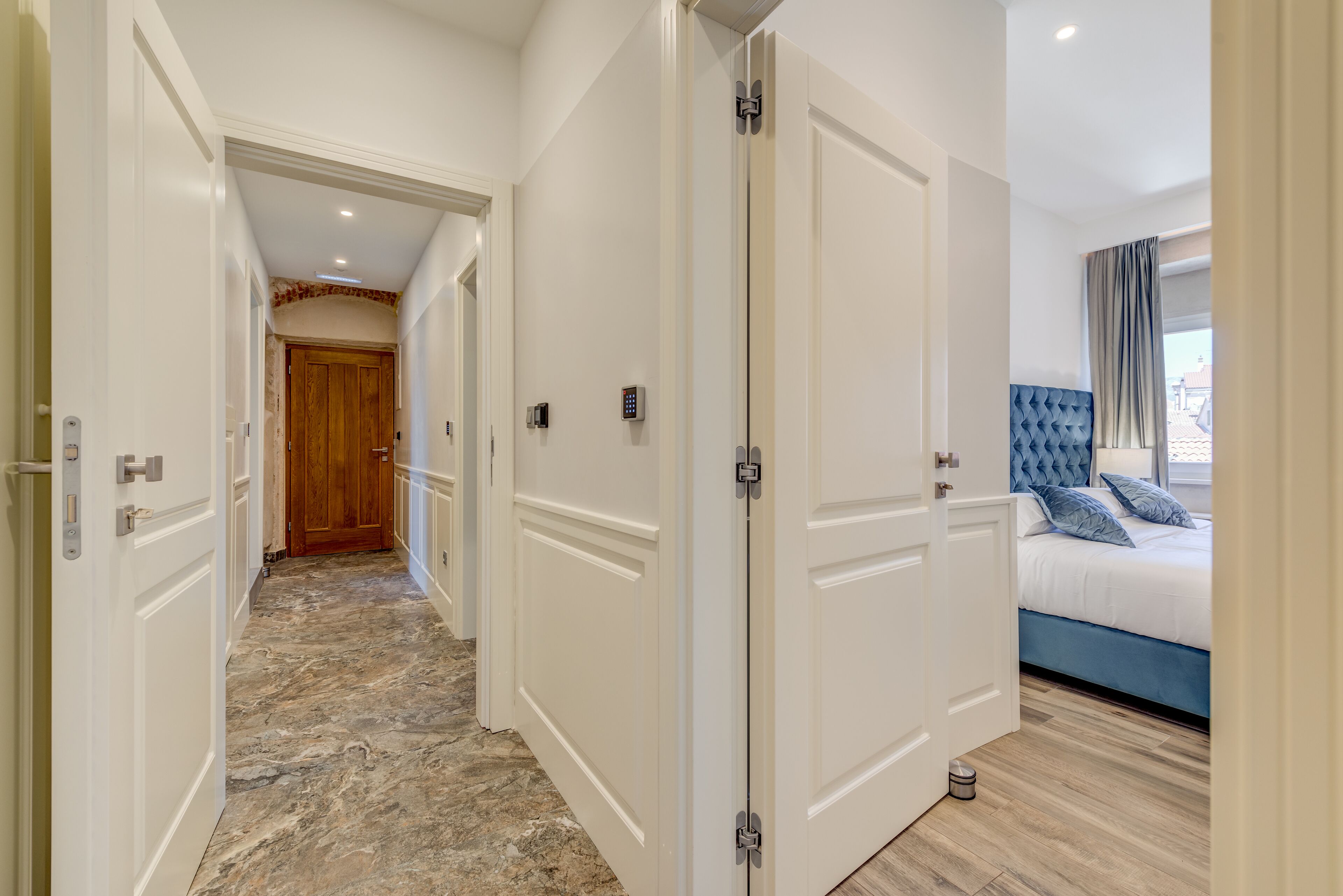 A hotel hallway with white doors, a marble floor, and a bedroom with a bed and blue headboard.