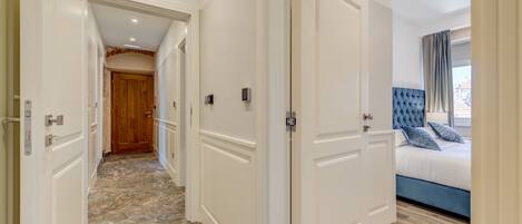 A hotel hallway with white doors, a marble floor, and a bedroom with a bed and blue headboard.
