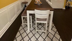 Een eetruimte met een donker houten tafel, witte stoelen en een kamerkleed met een patroon.