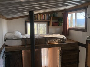 Un lit superposé avec une tête de lit, une fenêtre donnant sur des arbres, une bibliothèque remplie de livres et d’objets décoratifs, et un radiateur.