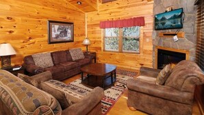 A cozy log cabin interior with a fireplace, a television mounted on the wall, and a comfortable seating area with a couch and armchairs.