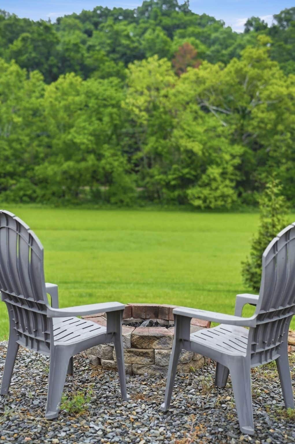 Zwei Adirondack-Stühle zeigen auf eine Feuerstelle aus Stein, im Hintergrund ein Rasen und Bäume.