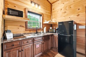 A compact kitchen with wooden walls, a black refrigerator, a microwave, a sink, and a window with blinds.