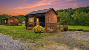 A rustic cabin with a porch, surrounded by a gravel driveway and grassy area, set against a backdrop of trees and a twilight sky. - Romantic Smoky Mountain Cabin | Hot Tub | Pet-Friendly (Sevierville)