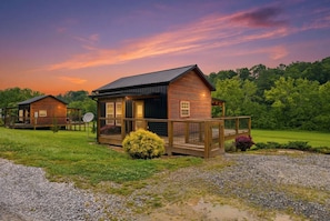 A rustic cabin with a porch, surrounded by a gravel driveway and grassy area, set against a backdrop of trees and a twilight sky. - Romantic Smoky Mountain Cabin | Hot Tub | Pet-Friendly (Sevierville)