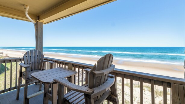 Eine Holzterrasse mit zwei Adirondack-Stühlen und einem Tisch mit Blick auf einen Strand.
