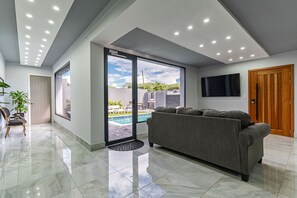 Een moderne woonkamer met een grijze bank, een aan de muur bevestigde televisie en een grote schuifpui naar een zwembad.