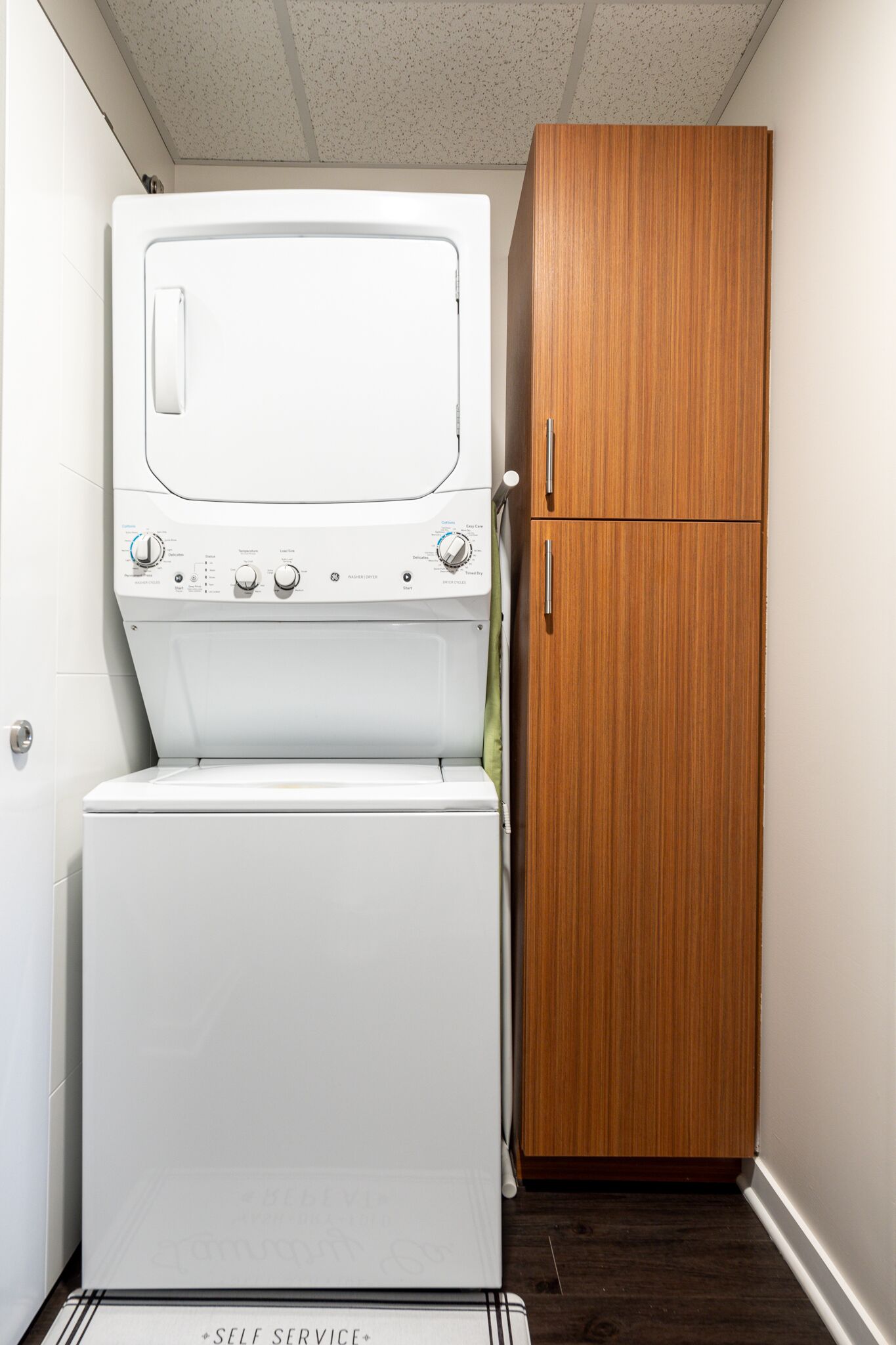 A compact laundry area with a washing machine and dryer stacked vertically, a wooden cabinet, and a door marked "SELF SERVICE".