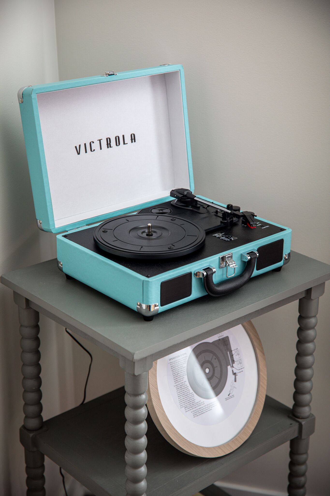 A turquoise suitcase with the word "VICTROLA" on the inside lid, placed on a wooden table with a turntable inside.