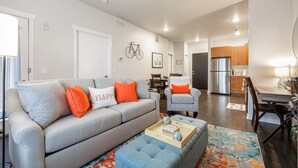 A modern living room with a grey sofa, orange pillows, and a blue ottoman. There is a dining area with a wooden table and chairs, and a kitchen in the background. - Cute Condo | TART Trail | Bikes | AC | Downtown | (Traverse City)