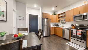 A modern kitchen with wooden cabinets, stainless steel appliances, and a dining area with a wooden table and chairs. - Cute Condo | TART Trail | Bikes | AC | Downtown | (Traverse City)