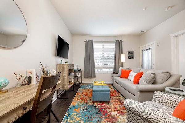 Bright living area w/ plush sofa, colorful rug & TV, flowing into a cozy dining space by the window.