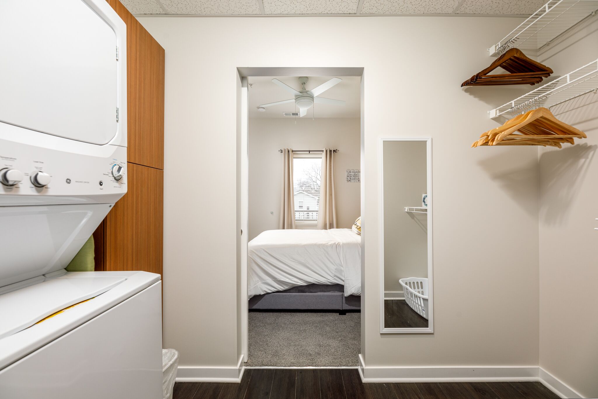 A laundry room with a washer and dryer, a bed with a gray mattress, a ceiling fan, and a mirror on the wall.