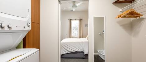 A laundry room with a washer and dryer, a bed with a gray mattress, a ceiling fan, and a mirror on the wall.