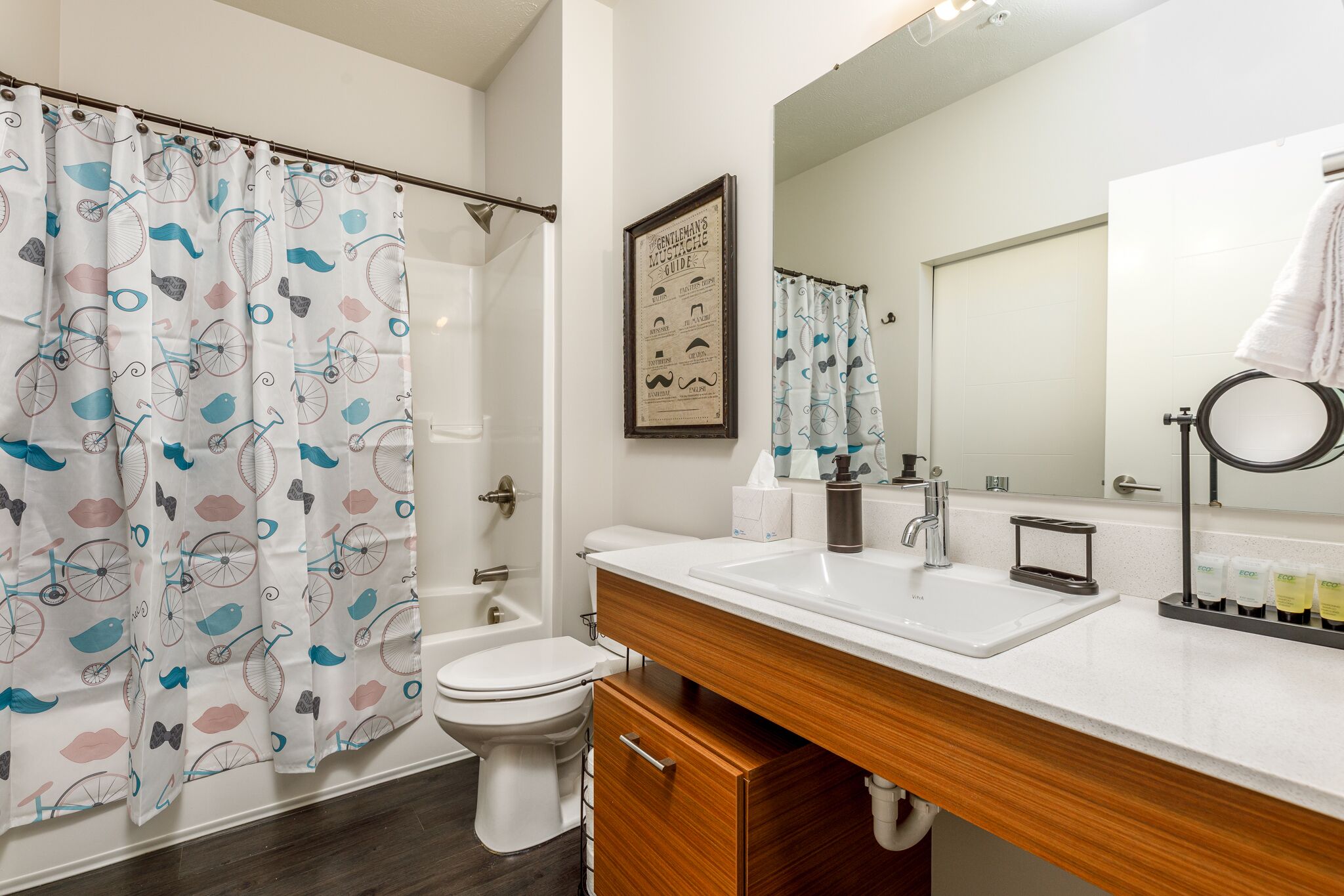 Une salle de bain moderne avec un rideau de douche orné de motifs de vélos et d’oiseaux, des toilettes, un lavabo avec un miroir et un guide encadré accroché au mur.