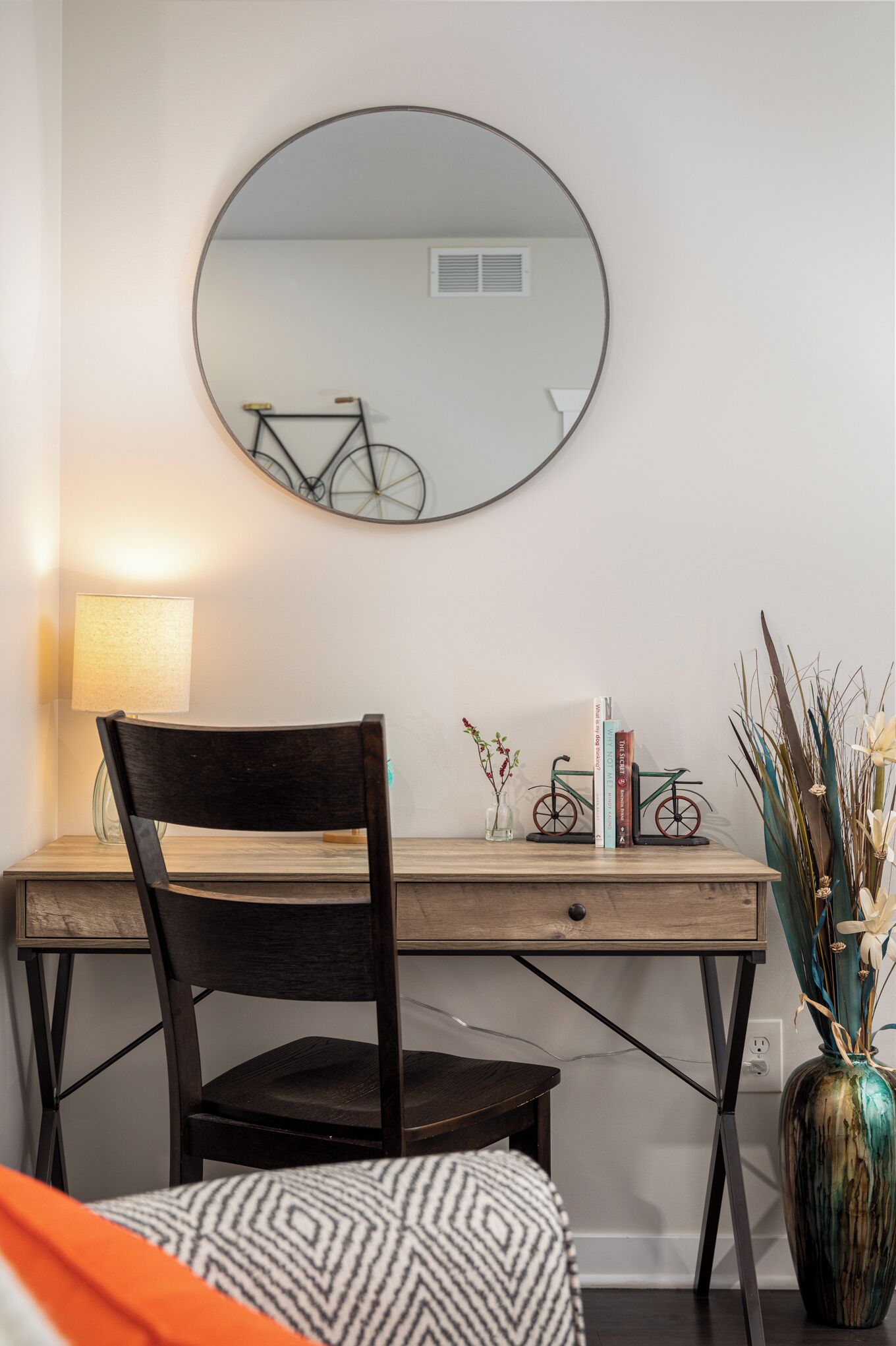 A room with a wooden desk, a chair, a round mirror, a lamp, a vase with plants, and a bicycle decoration.