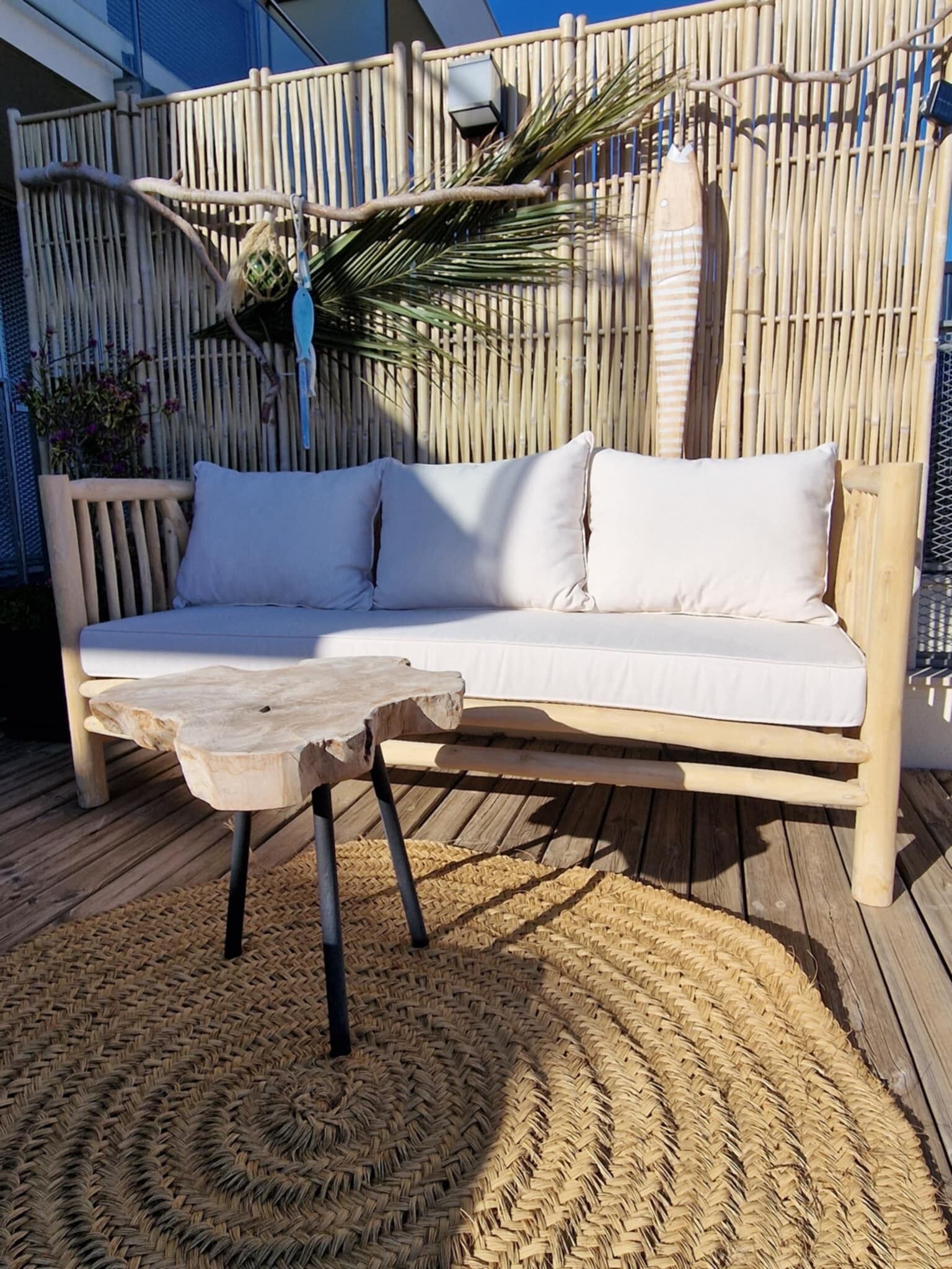 Una terraza de madera con alfombra tejida, un banco de madera y un sofá blanco con cojines junto a una valla de bambú.