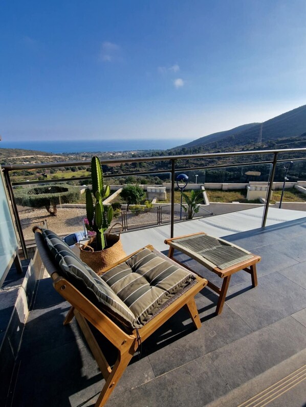 A balcony with two wooden lounge chairs, a potted cactus, and a view of the sea and hills.