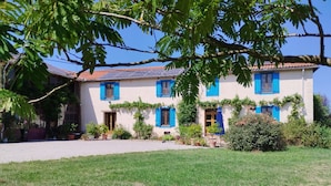A two-story house with a white exterior and blue shutters, surrounded by a well-maintained lawn and various plants. - Former farm, very quiet location, lots of space (Libaros)