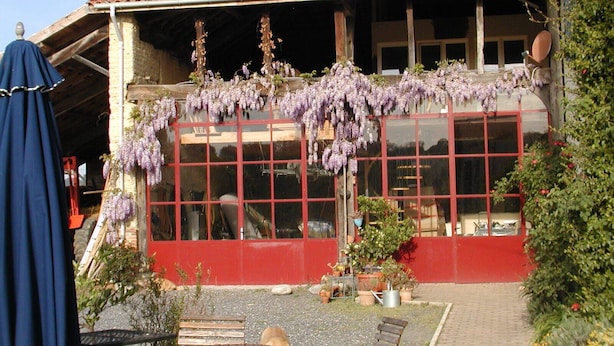 A building with a red facade, large windows, and hanging purple flowers. There are outdoor seating areas with tables and chairs. - Former farm, very quiet location, lots of space (Libaros)
