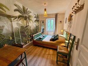 Un dormitorio con cama, una silla, una mesa, un ventanal y un mural temático tropical.