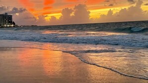 Una playa con olas al atardecer, edificios al fondo y un cielo despejado.