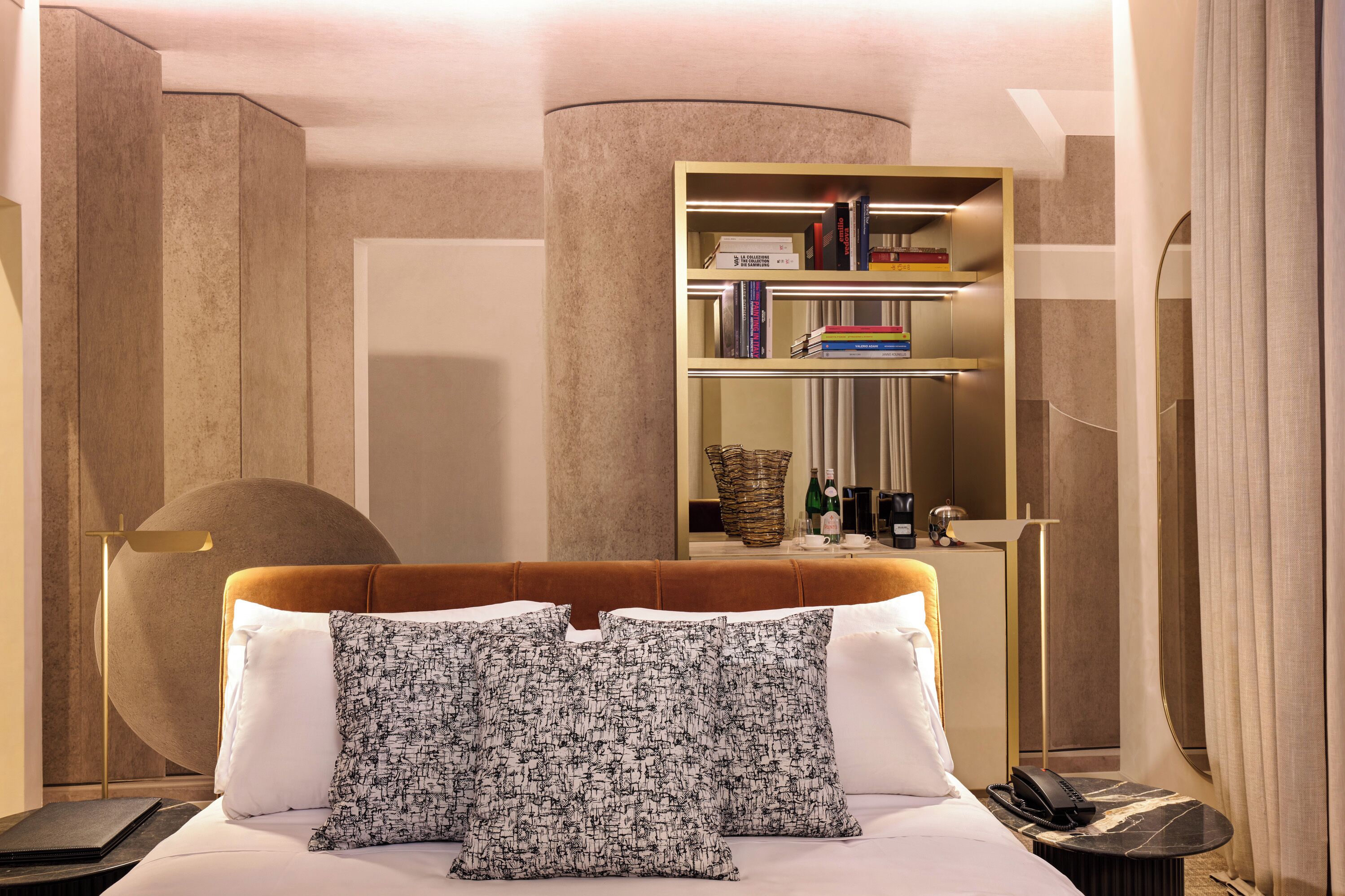 A hotel room with a bed, a bedside table with a phone, a bookshelf, and a lamp.