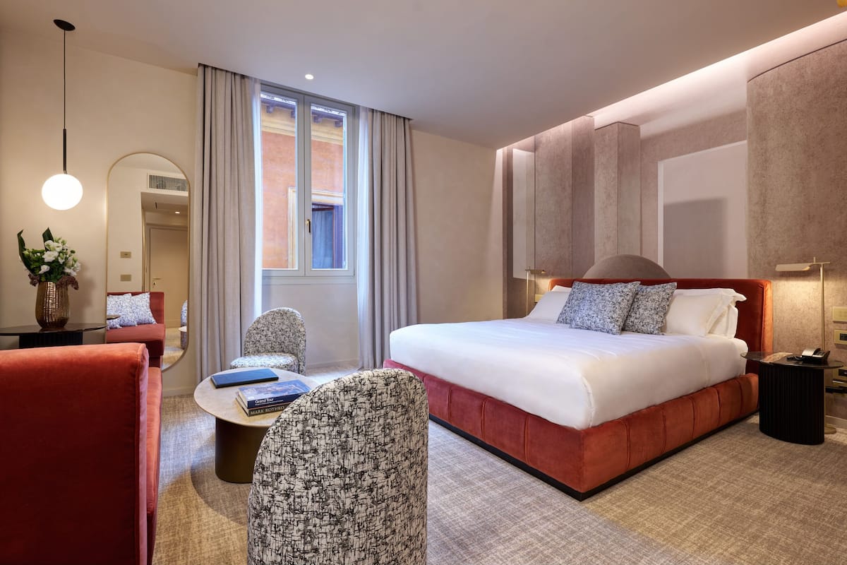 A modern hotel room with a large bed, a seating area with a round table, and a view of the outside through a window.