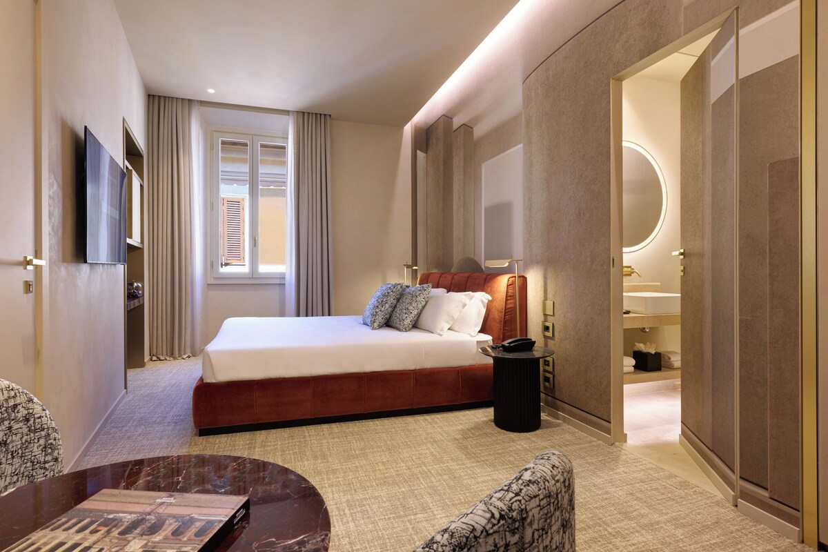 A hotel room with a bed, a TV, a bedside table, a chair, a desk, and a bathroom with a mirror and sink.