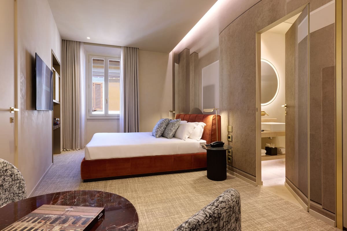 A hotel room with a bed, a TV, a bedside table, a chair, a desk, and a bathroom with a mirror and sink.