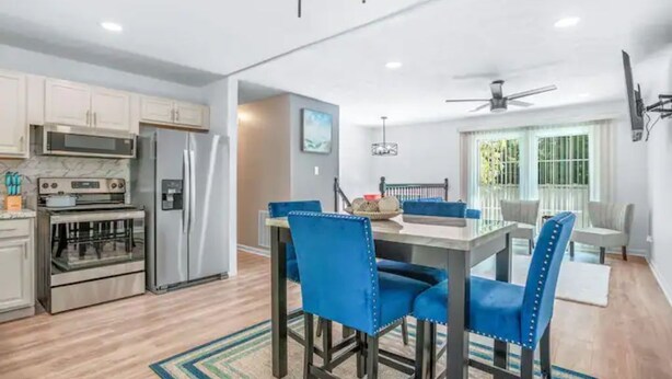 Una cocina moderna con electrodomésticos de acero inoxidable, un comedor con sillas tapizadas en azul y un ventilador de techo.