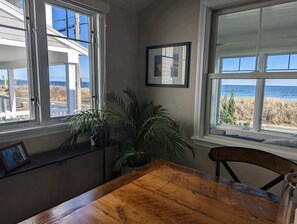A room with a wooden table and chairs, a potted plant, and a view of the beach through large windows.