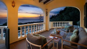 A balcony with a view of the sea at sunset, furnished with a sofa, chairs, and a table with books and a bottle.