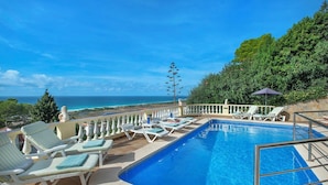 A pool area with lounge chairs, a diving board, and a view of the ocean.