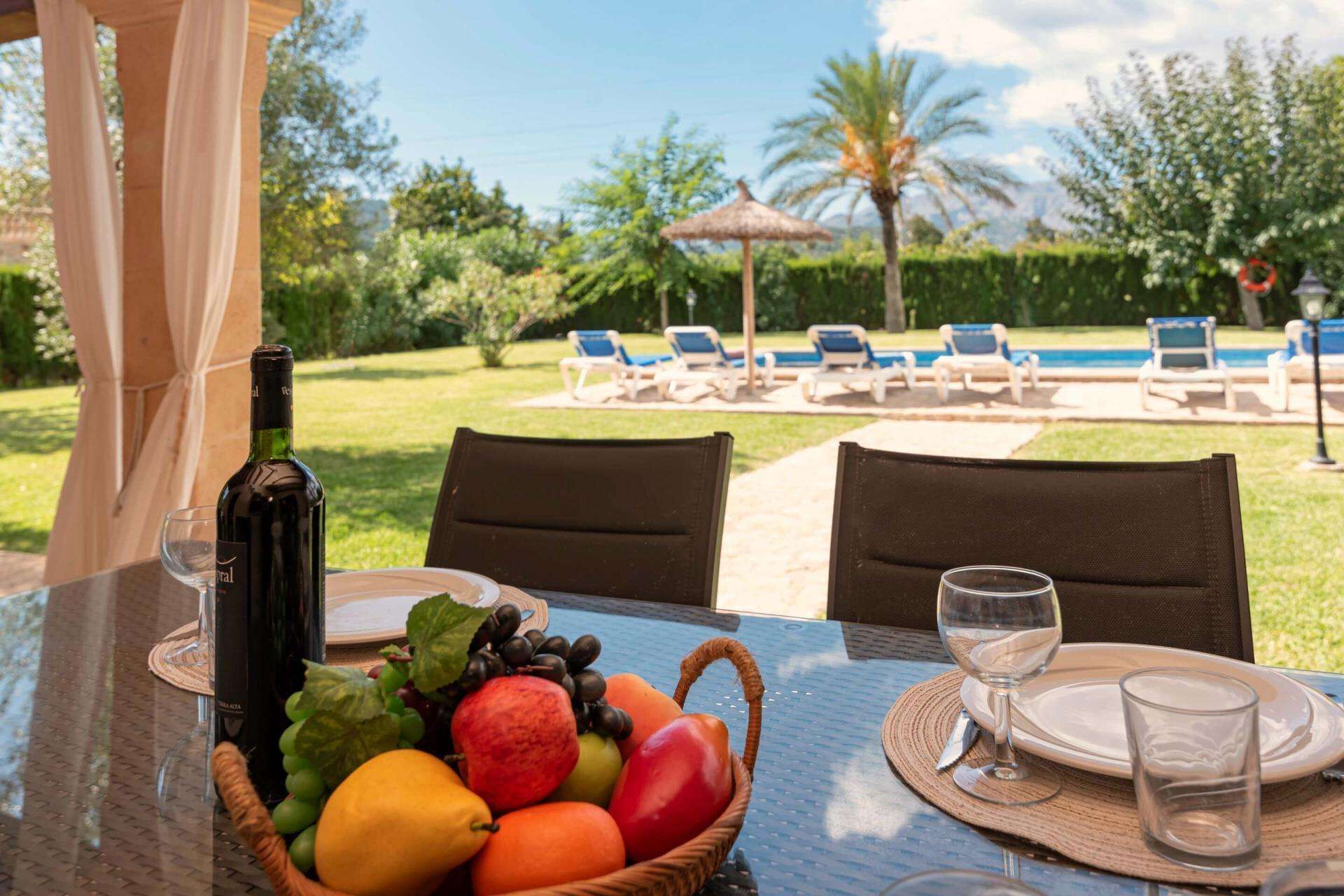 A table with a bottle of wine, a basket of fruit, and a view of a pool area with lounge chairs and a palm tree.