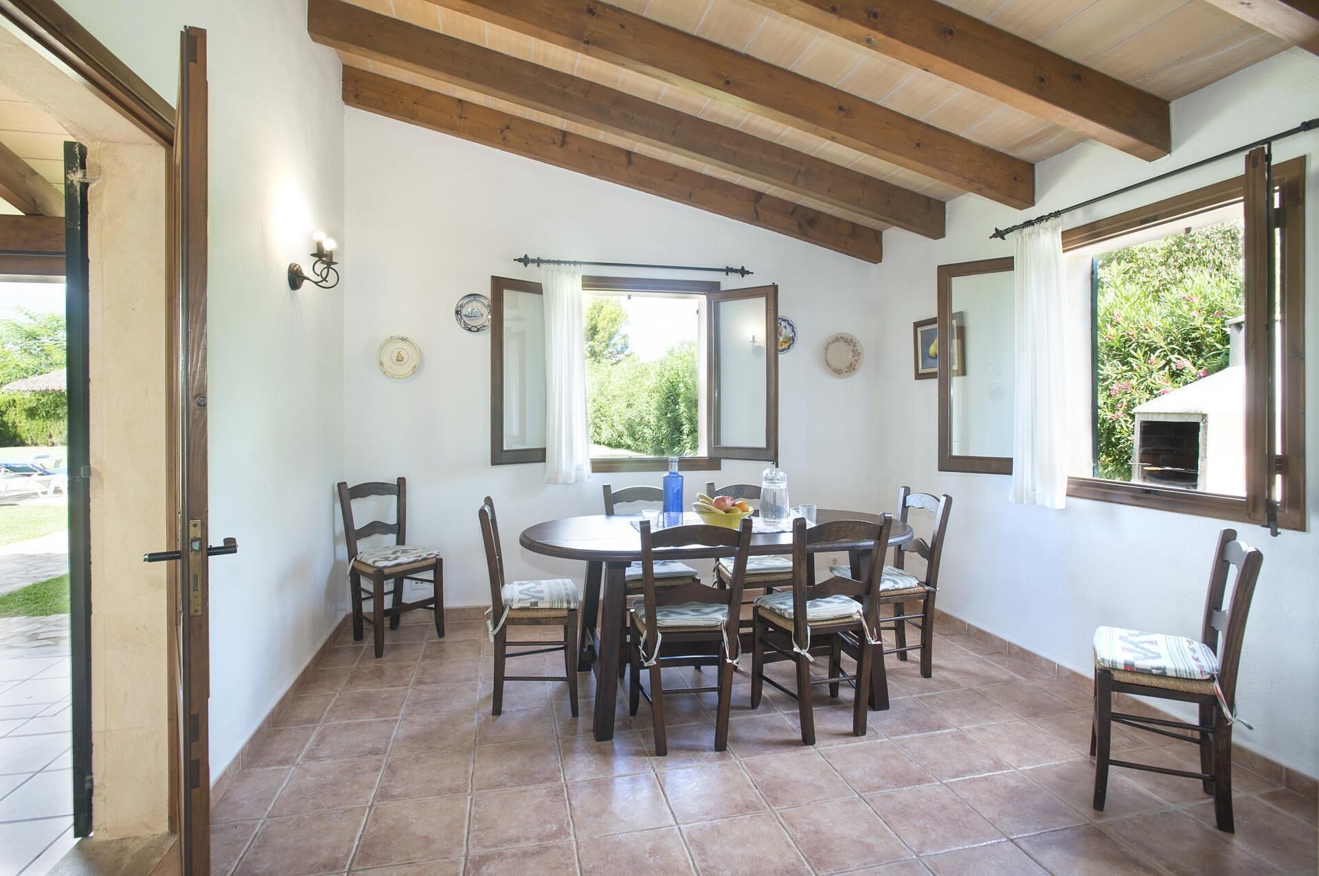 Un comedor con una mesa redonda y seis sillas, vigas de madera en el techo y una ventana con vistas a la vegetación.