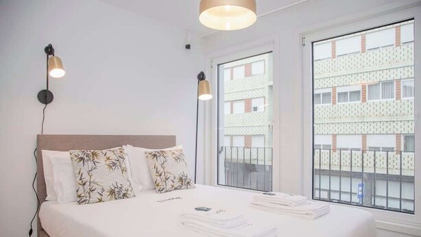 Ein ordentlich bezogenes Bett mit Blumenkissen, eine Nachttischlampe und Blick auf ein Gebäude durch ein Fenster.