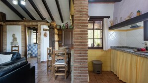 A rustic interior with exposed brick walls, a stone fireplace, a dining area with wooden chairs, and a kitchen with a yellow countertop.