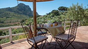 An outdoor dining area with a table set for two, surrounded by metal chairs. A thatched umbrella provides shade. The setting overlooks a lush valley with terraced fields and a prominent mountain in the distance.